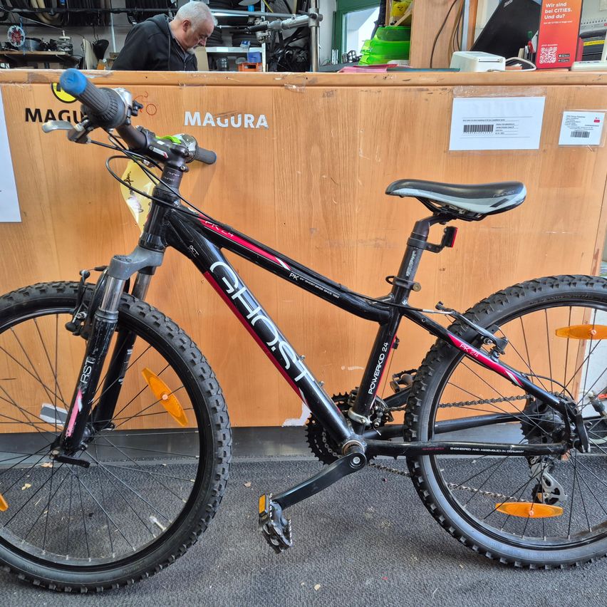 Ein schwarzes und pinkes Fahrrad, Modell GHOST POWERBIRD 24, steht vor einem hölzernen Tresen mit der Aufschrift Magura. Ein älterer Mann sitzt hinter dem Tresen.