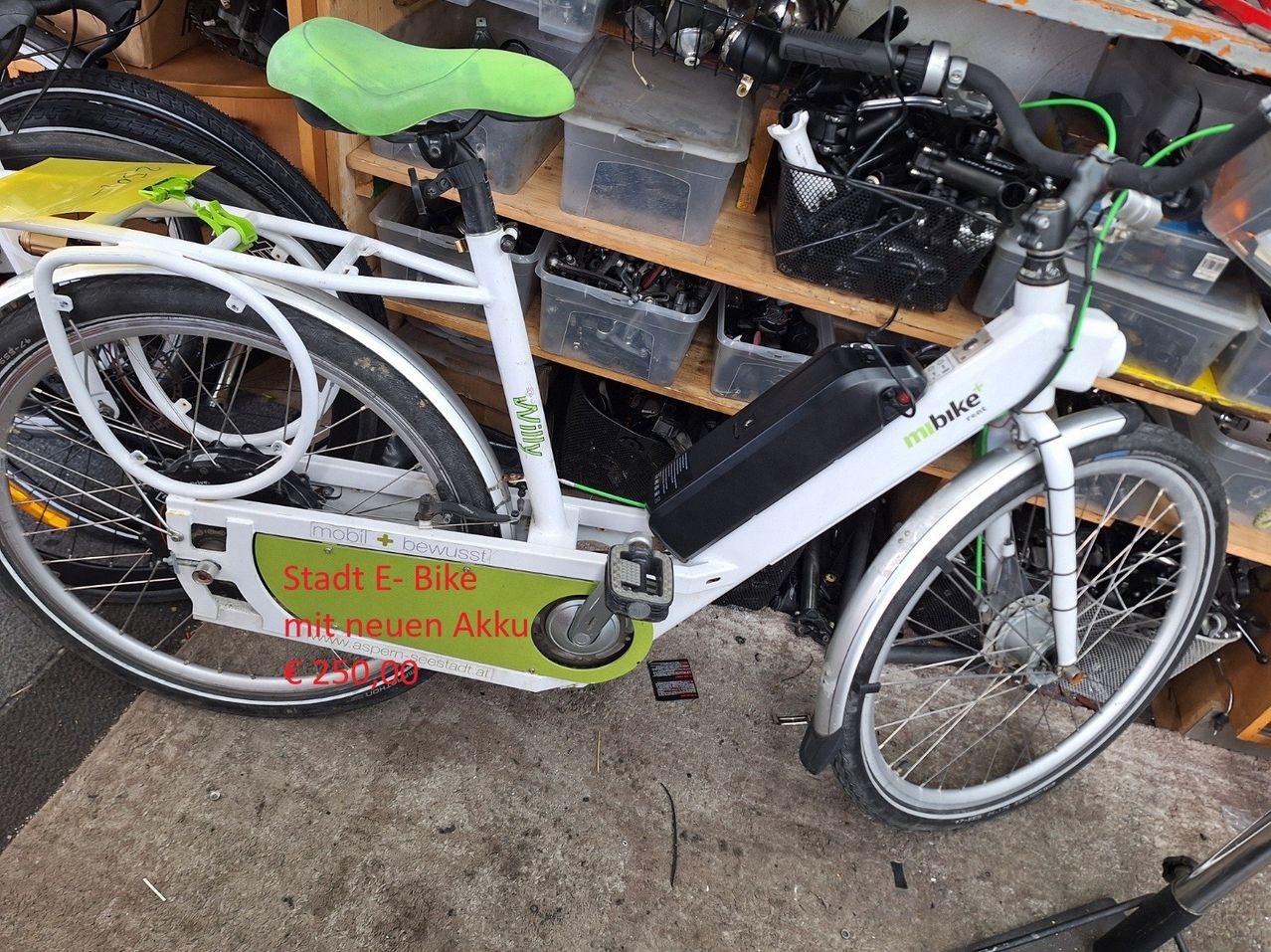 Ein weißes und grünes E-Bike mit grünem Sattel und Akku steht auf dem Boden in der Nähe von Regalen mit Aufbewahrungsboxen.