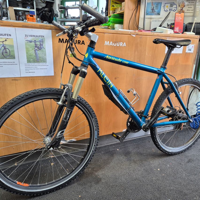 Ein blaues Fahrrad, Modell Attitude Comp, steht in einer Werkstatt. Es hat einen schwarzen Sattel, eine Wasserflasche und silberne Lenker. Dahinter befindet sich ein hölzerner Schreibtisch mit einem Schild, auf dem MAURA steht.
