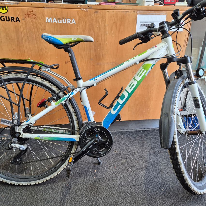 Ein weiß-blaues Cube-Fahrrad mit schwarzem Sattel steht vor einem hölzernen Tresen. Das Fahrrad hat einen Gepäckträger und eine Kettenschutz.
