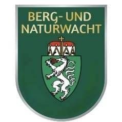 Ein grüner Schild zeigt ein Wappen mit einem weißen Löwen, der ein rotes Objekt hält, umgeben von einem roten und goldenen Kreis. Über dem Schild befindet sich ein Kreuz auf rotem Hintergrund. Die Worte 'BERG-UND NATURWACH' sind in Fettschrift über dem Schild geschrieben.