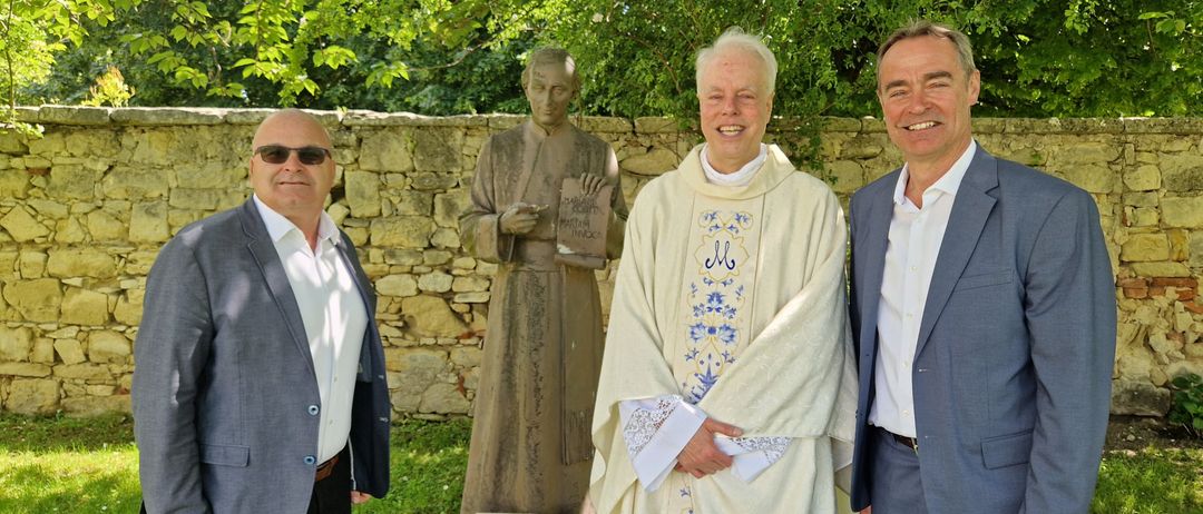 Drei Männer stehen vor einer Statue und einer Steinmauer und lächeln. Einer hält ein Buch, ein anderer trägt eine weiße Robe mit blauen Blumen und der dritte trägt einen grauen Anzug.