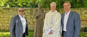 Drei Männer stehen vor einer Statue und einer Steinmauer und lächeln. Einer hält ein Buch, ein anderer trägt eine weiße Robe mit blauen Blumen und der dritte trägt einen grauen Anzug.