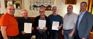 Fünf Männer stehen in Innenräumen und halten Urkunden in der Hand. Sie tragen alle eine Brille, Schuhe und Gürtel. Der Raum ist mit einem Vorhang, einem Regal und anderen Gegenständen dekoriert.