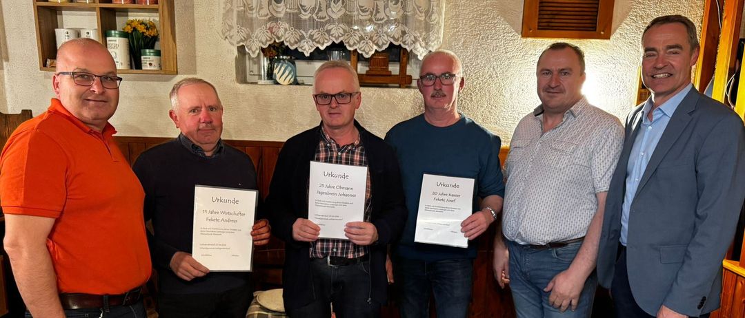 Five men stand indoors holding certificates. They all wear glasses, shoes, and belts. The room is decorated with a curtain, shelf, and other items.