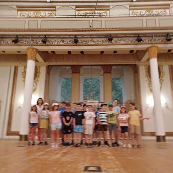 Eine Gruppe von Kindern und Erwachsenen steht in einer Reihe in einem großen Saal mit aufwendigen Säulen und einer Bühne.