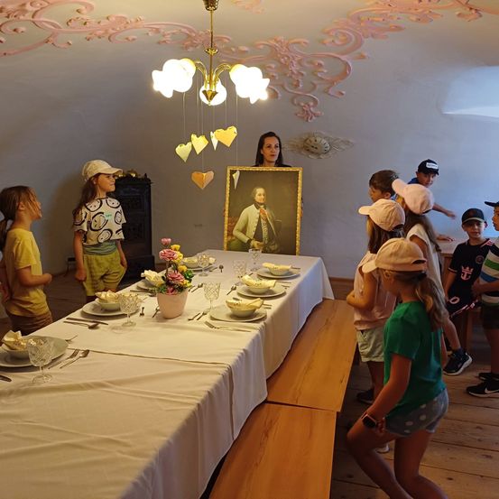 Kinder versammeln sich um einen Tisch, eine Frau steht mit einem Porträt in den Händen. Sie tragen Hüte und Turnschuhe.
