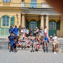 Eine Gruppe von Kindern und Erwachsenen posiert vor einem gelben Gebäude mit grünen Fensterläden und einem Balkon.