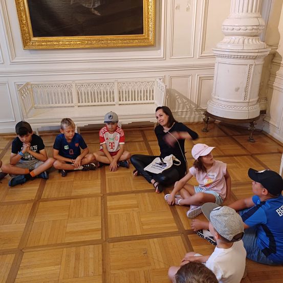 Eine Gruppe von Kindern sitzt auf dem Boden vor einer Frau, die mit ihnen spricht. Der Raum hat weiße Wände und Holzböden. Es gibt eine weiße Bank und eine weiße Säule im Raum.