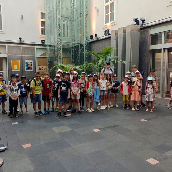 Eine Gruppe von Kindern und Erwachsenen steht vor einem Gebäude. Die Kinder tragen Hüte und Turnschuhe, und einige von ihnen haben Rucksäcke dabei.