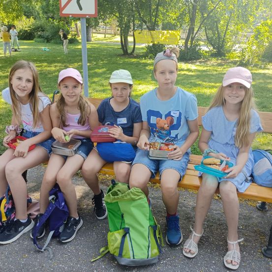 Fünf Mädchen sitzen auf einer Bank, lächeln und halten Lunchboxen. Sie tragen Mützen, Turnschuhe und Sandalen. Ein Rucksack liegt auf dem Boden. Die Bank steht draußen in der Nähe einer Rasenfläche, und es gibt Bäume und ein Schild im Hintergrund.
