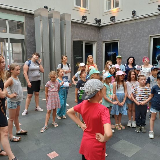 Eine Gruppe von Kindern und Erwachsenen versammelt sich in einem Innenhof, angeführt von einer Frau. Der Innenhof hat graue Wände und einen gefliesten Boden.