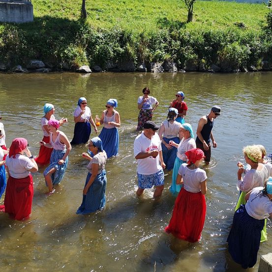 Eine Gruppe von Menschen in traditioneller Kleidung steht in einem Fluss, einige halten Gläser und Flaschen. Sie scheinen an einer Flusssegnungszeremonie teilzunehmen.
