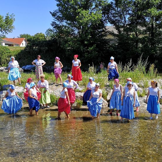 Eine Gruppe von Frauen in farbenfrohen Kleidern und Kopfbändern steht in einem flachen Fluss, einige mit Sonnenbrille. Im Hintergrund befinden sich Bäume und Häuser.