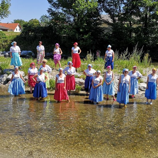 Eine Gruppe von Frauen steht in einem Fluss, trägt traditionelle Kleidung und Sonnenbrillen. Sie sind in einer Reihe aufgestellt, wobei eine Person vor der anderen steht. Der Fluss ist von Felsen und Pflanzen umgeben. In der Ferne befindet sich ein Haus mit einem roten Dach.