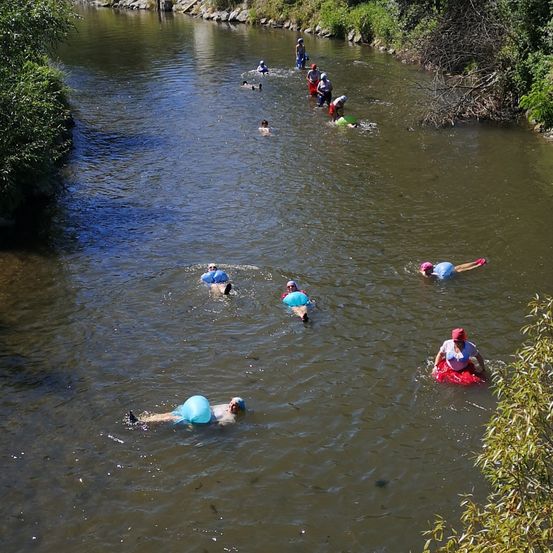 Mehrere Menschen treiben mit Helmen und Schwimmkappen auf einem Fluss. Einige liegen auf aufblasbaren Geräten, während andere schwimmen.