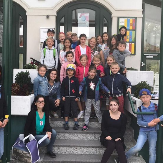 Eine Gruppe von Kindern und zwei Frauen steht vor einem Gebäude mit grünen Türen. Einige Kinder tragen Rucksäcke. Ein grünes Banner mit dem Wort 'BU' hängt an der Wand.
