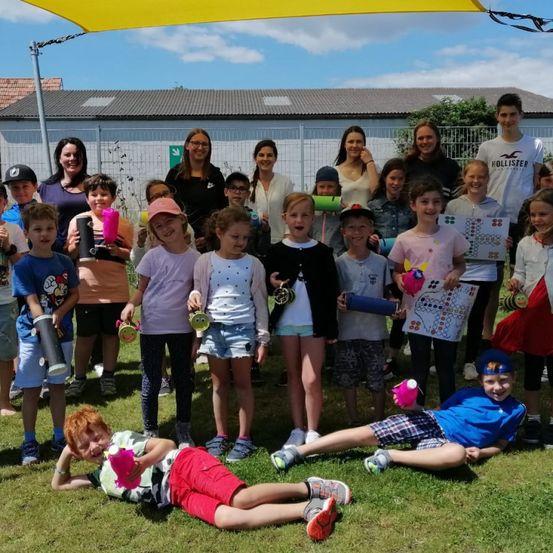 Gruppe von Kindern und Erwachsenen, die auf einem Rasen stehen und liegen, verschiedene Bastelarbeiten in den Händen halten, ein Kind hält einen rosa Becher, ein anderes ein Brettspiel, dahinter ein weißer Zaun und ein Gebäude