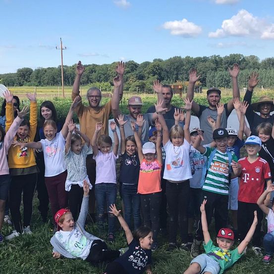 Eine Gruppe von Kindern und Erwachsenen posiert für ein Foto auf einer Wiese mit einem Zaun und Bäumen im Hintergrund.