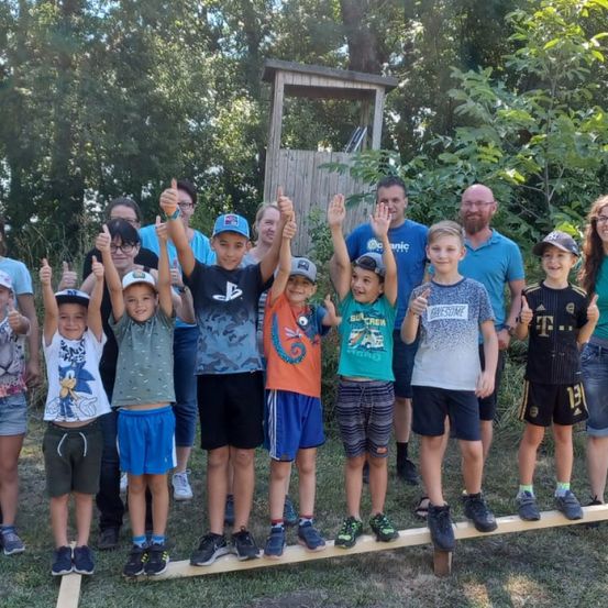 Eine Gruppe von Kindern und Erwachsenen steht draußen auf einem Holzbrett, lächelt und gibt Daumen hoch.