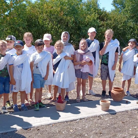 Eine Gruppe von Kindern, die in weißen Togen gekleidet sind, posiert für ein Foto auf einem Betonpfad, mit Tonschüsseln auf dem Boden und Bäumen im Hintergrund.