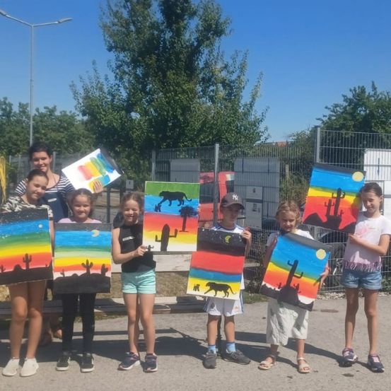 Eine Gruppe junger Mädchen und einer Frau, die Gemälde halten, steht in einer Reihe vor einem Zaun.