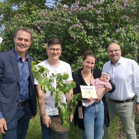 Drei Männer und eine Frau stehen draußen mit einem Topfpflanzen. Ein Mann hält ein Zertifikat. Die Frau hält ein Baby. Sie alle lächeln.