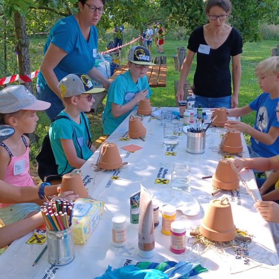 Mehrere Kinder bemalen Tontöpfe, während zwei Frauen zusehen. Im Hintergrund stehen und gehen Personen.