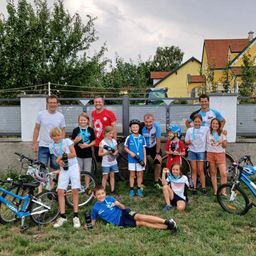 Gruppenfoto von Kindern und Erwachsenen, die in einem Grasbereich stehen und sitzen, alle tragen Radsporttrikots und Medaillen. Fahrräder und Zäune sind im Hintergrund.