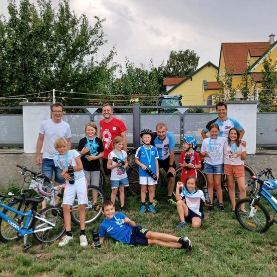 Gruppenfoto von Kindern und Erwachsenen, die in einem Grasbereich stehen und sitzen, alle tragen Radsporttrikots und Medaillen. Fahrräder und Zäune sind im Hintergrund.