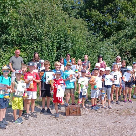 Eine Gruppe von Kindern, Erwachsenen und älteren Menschen steht in einer Reihe auf einer Straße. Sie halten Pokale, Urkunden und Flaschen. Ein Holzkasten liegt auf dem Boden.
