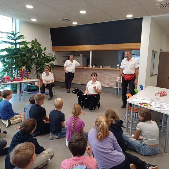 Eine Gruppe von Kindern sitzt auf dem Boden und beobachtet eine Frau mit einem schwarzen Hund in einem Raum. Hinter ihr stehen zwei Männer, einer mit einem roten Gürtel. Der Raum hat Tische und Stühle, Pflanzen und eine Theke.