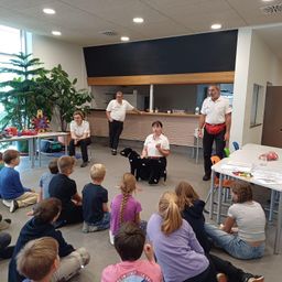 Eine Gruppe von Kindern sitzt auf dem Boden und beobachtet eine Frau mit einem schwarzen Hund in einem Raum. Hinter ihr stehen zwei Männer, einer mit einem roten Gürtel. Der Raum hat Tische und Stühle, Pflanzen und eine Theke.