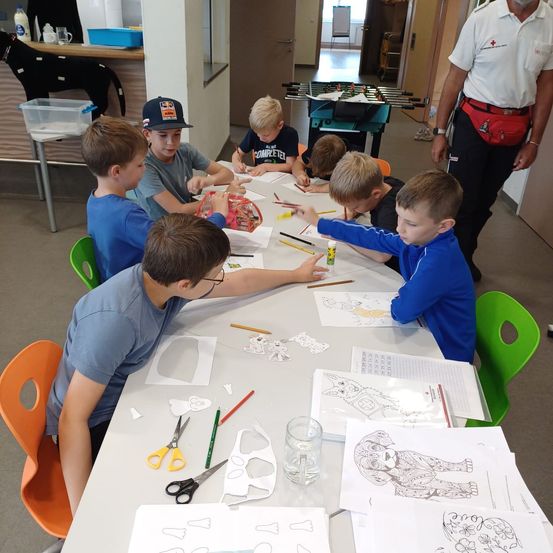 Eine Gruppe von Jungen sitzt um einen Tisch herum und schneidet und zeichnet Papiere mit Scheren. Sie sind in einem Raum mit Tischen, Stühlen und einer Hundefigur. Ein erwachsener Mann steht in der Nähe.