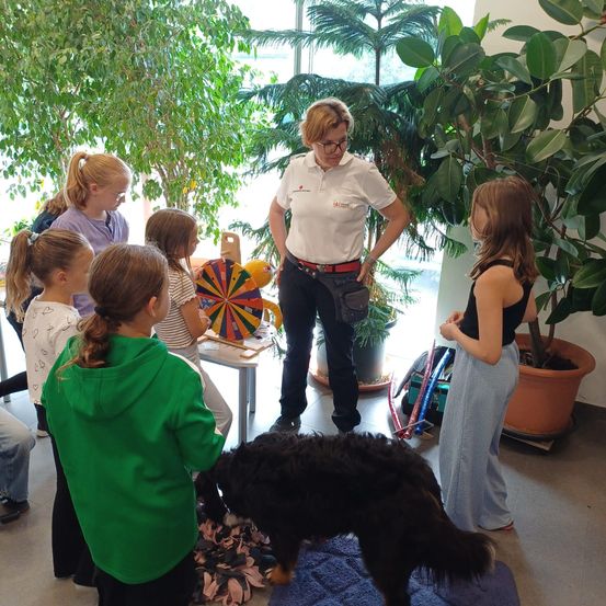 Eine Frau mit Brille und Gürtel erklärt etwas einer Gruppe von Mädchen in einem Raum mit einem Hund und großen Pflanzen.