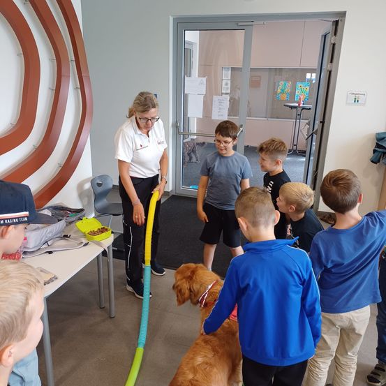 Eine Frau und mehrere Kinder stehen um einen Hund herum. Die Frau hält ein Spielzeug, und die Kinder beobachten, wie der Hund steht und mit ihnen interagiert. Ein Tisch mit Snacks steht hinter ihnen, und eine Tür führt in einen anderen Raum.