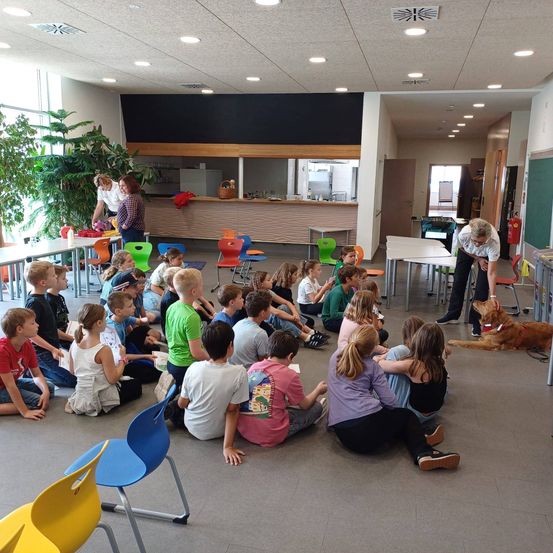 Eine Gruppe von Kindern sitzt in einem Klassenzimmer in einem Kreis mit einem Hund in der Nähe. Ein Erwachsener steht in der Nähe und zwei weitere Erwachsene sind an einer Theke.