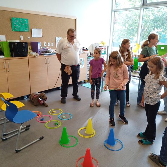 Ein Mann mit Brille trainiert einen Hund in einem Raum, in dem Kinder mit farbigen Kegeln und Ringen auf dem Boden spielen.