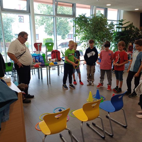 Eine Gruppe junger Jungen beteiligt sich in einem Raum mit Stühlen und Hula-Hoops an einem Spiel, überwacht von einem Mann mit Brille.