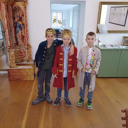 Drei Jungen stehen zusammen auf einem Holzboden, gekleidet in mittelalterliche Kleidung. Einer trägt einen roten Mantel mit Goldbesatz, ein anderer ein blaues Jackett mit Krone, und der dritte ein weißes Shirt mit buntem Aufdruck. Hinter ihnen steht eine Holzstruktur und ein Spiegel.