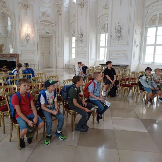 Mehrere Kinder mit Rucksäcken sitzen in Stühlen in einem eleganten Saal mit hohen Decken und kunstvollen Verzierungen.