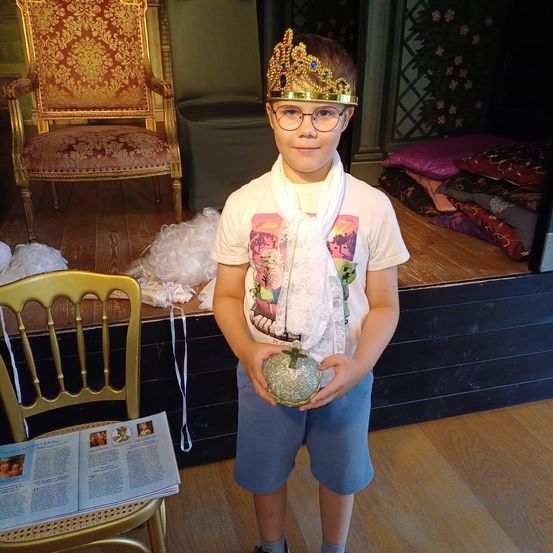 Ein Junge mit Krone und Ball steht auf der Bühne mit einem Stuhl und Kissen im Hintergrund.