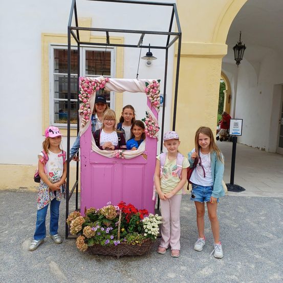 Eine Gruppe junger Mädchen mit zwei Erwachsenen posiert vor einer pinkfarbenen Tür, die mit Blumen geschmückt ist. Der Ort ist im Freien in der Nähe eines Gebäudes mit einer Lampe.