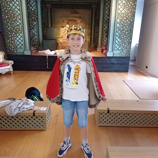 Ein junger Junge ist in einem roten Umhang und einer Krone gekleidet und steht auf der Bühne eines Theaters mit einem Hintergrund aus einem goldenen Thron und Blumentapete.