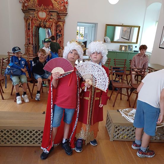 Zwei Kinder in historischen königlichen Kostümen stehen in einem Raum und halten Fächer. Hinter ihnen sitzen mehrere Kinder auf Stühlen, andere stehen herum. Im Hintergrund befinden sich ein Spiegel und ein Schreibtisch.