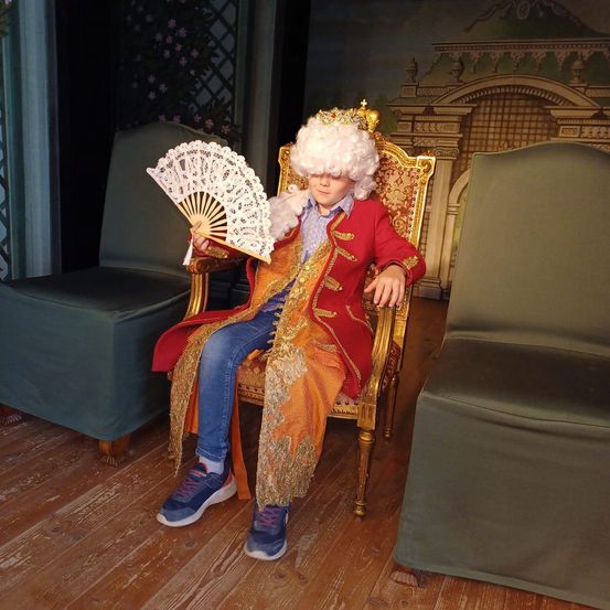 Ein Kind in einem königlichen Kostüm mit Krone, das einen Spitzenfächer hält, sitzt auf einem goldenen Stuhl, umgeben von grünen Stühlen und einem Holzboden.