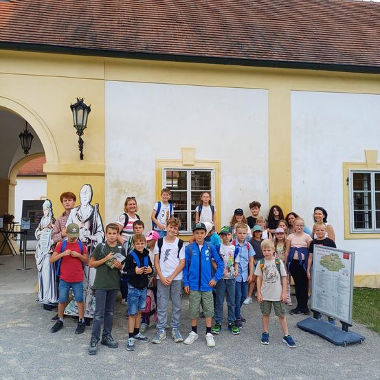 Eine Gruppe von Kindern und Erwachsenen steht vor einem historischen Gebäude mit Statuen. Einige tragen Hüte und Rucksäcke. Das Gebäude hat gewölbte Eingänge, Fenster und Lampen.