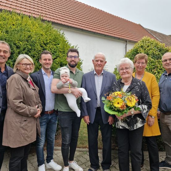 Eine Gruppe von Menschen, darunter ein Baby, steht vor einem Haus mit einem roten Dach. Die Menschen sind in verschiedene Kleidungsstücke gekleidet, und einige von ihnen halten Blumen. Der Mann in der Mitte hält ein Baby und lächelt.