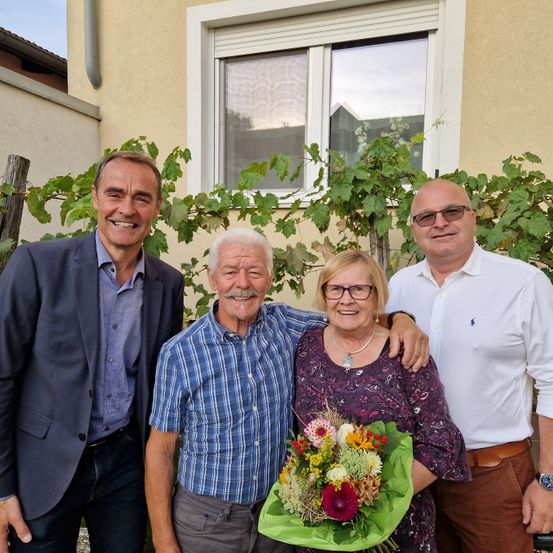 Vier Personen stehen vor einem Haus mit einem Fenster. Sie alle haben fröhliche Gesichtsausdrücke. Der Mann links trägt einen blauen Blazer, und der Mann rechts trägt ein weißes Hemd. Die Frau in der Mitte hält einen Blumenstrauß.