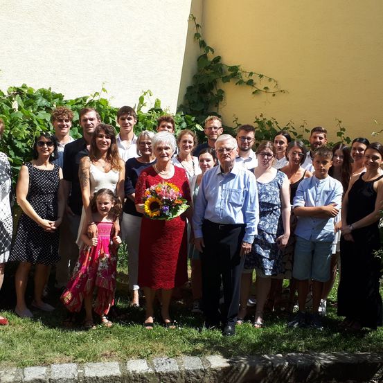 Eine Familie versammelt sich im Freien, wobei ein älteres Paar vorne und in der Mitte steht. Sie halten Blumen. Dahinter stehen Menschen jeden Alters in Reihen, wobei einige lächeln. Der Hintergrund zeigt eine gelbe Wand mit Kletterpflanzen.